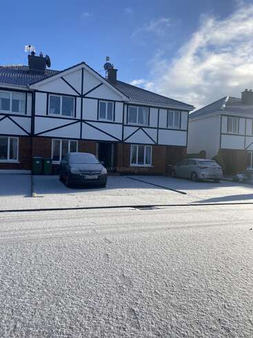 A row of modern houses with parked cars, all covered in a light layer of snow under a bright blue sky with scattered clouds, winter morning ambiance.