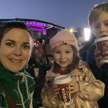 A woman and two children smile at an outdoor evening event, holding Costa coffee cups. Purple stage lights glow in the background with a crowd nearby.