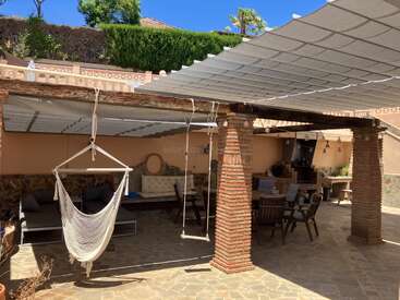 Esta imagen muestra un acogedor patio exterior con columnas de ladrillo, un toldo a la sombra, una hamaca, sofás, una mesa de comedor con sillas y una barbacoa.