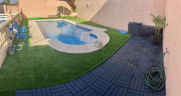 En el patio trasero hay una pequeña piscina enterrada, césped artificial, sillas plegables azules, una terraza de madera, una manguera de jardín, una maceta y paredes de color beige.