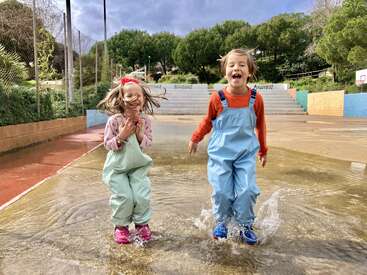 Dos niños alegres con monos impermeables saltan y chapotean en un charco. Las risas llenan el aire, rodeados de verdes árboles y coloridos escalones de parque infantil.