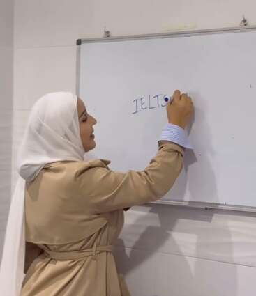 Uma mulher com casaco bege e lenço branco na cabeça escreve "IELTS" em um quadro branco em uma sala de aula, preparando-se para uma aula de inglês ou para um exame.