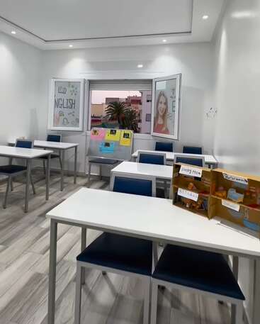 Esta imagem mostra uma sala de aula bem iluminada e arrumada, com carteiras brancas, cadeiras azuis, pôsteres educativos, uma janela e um modelo de casa de papelão com os cômodos identificados.