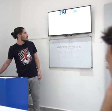 Um homem com cabelos longos em um coque está ao lado de um quadro branco e de uma tela de TV, parecendo explicar ou ensinar algo em uma sala de aula ou escritório.