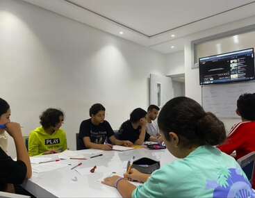 Um grupo de alunos está sentado ao redor de uma mesa branca em uma sala de aula moderna, fazendo anotações, assistindo a uma tela e ouvindo atentamente a uma aula.