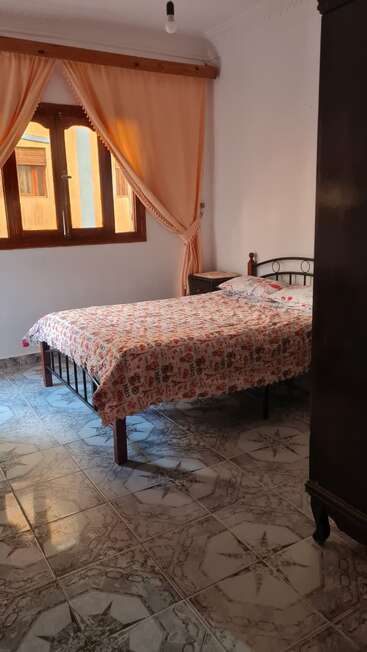 Um quarto aconchegante com piso de azulejos, estrutura de ferro da cama, colcha floral, cortinas pêssego cobrindo uma janela de madeira, uma mesa de cabeceira e luz solar natural preenchendo o espaço.