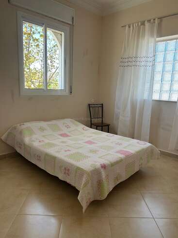 A simple bedroom with a single bed covered in a pastel patchwork quilt, two windows, white curtains, a tiled floor, and a black metal chair.