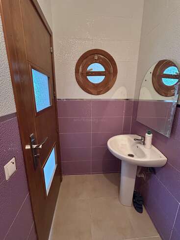 This small bathroom features purple wall tiles, a wooden door with frosted glass panels, a porthole-style window, a pedestal sink, mirror, and hand soap.