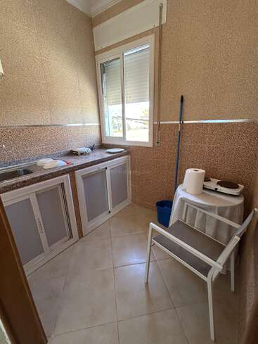 A small kitchen space with beige tiled walls, countertop, sink, window, white chair, mop, round table with hotplate, paper towel, blue bucket, and plates.