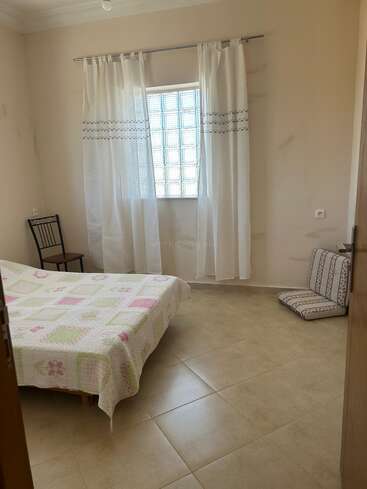 A simple, tidy bedroom with a single bed covered by a patterned quilt, a chair beside it, light curtains, tiled floor, and a floor cushion.