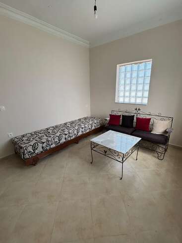 This room features a simple, minimalistic design with a patterned bench, metal-framed sofa with cushions, glass coffee table, cream walls, tiled floor, and window.