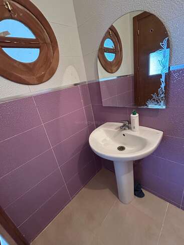 This small bathroom features a white pedestal sink, a mirror, purple-tiled walls, a round porthole window, beige floor tiles, and a single soap dispenser.