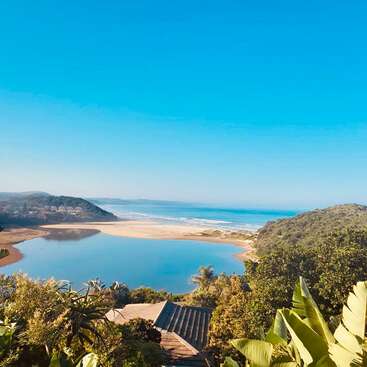 Une vue imprenable sur la côte, avec une végétation luxuriante, un lagon bleu paisible, une plage de sable, des collines ondulantes et un ciel dégagé, compose un paysage naturel serein et accueillant.