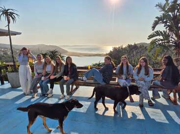 Nine young women sit on a bench overlooking a scenic landscape with water, hills, and sunlight. Two black dogs stand on the blue-painted patio.