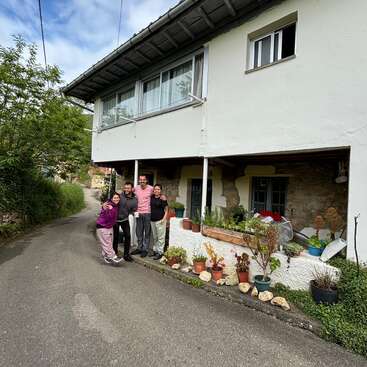 L'image représente une maison blanche à deux étages avec un groupe de quatre personnes debout devant, entourées de plantes en pot et d'une route pavée menant à la maison.
