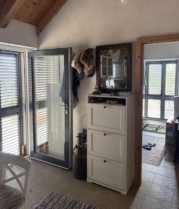Uma entrada aconchegante com uma porta de vidro aberta, chapéus pendurados na parede, um armário branco, um espelho, a luz do sol entrando e um cômodo visível ao fundo.