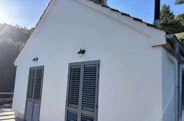 Essa imagem mostra uma casinha branca com telhado de telhas vermelhas, duas janelas com venezianas, luminárias de parede externas e colinas arborizadas visíveis ao fundo.