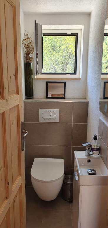 Esta é uma pequena casa de banho minimalista com vaso sanitário, pia compacta, sabonete, lixeira, janela, vaso decorativo, espelho e porta de madeira. Luz natural abundante.