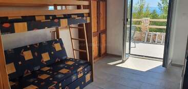 Esta imagem mostra um quarto bem iluminado com beliches, roupa de cama estampada, um guarda-roupa de madeira, piso de azulejo e uma porta aberta que dá para uma varanda com uma vista panorâmica.