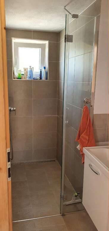 Um banheiro moderno com azulejos bege, porta de vidro no box, toalha laranja, uma janelinha com produtos de higiene pessoal, chuveiro de mão e um armário branco embaixo da pia.