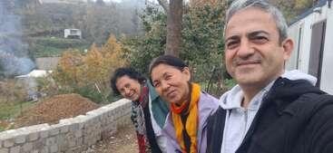 Drei Leute, zwei Frauen und ein Mann, lächeln draußen in einer ländlichen Umgebung mit Herbstbäumen, einer Steinmauer, Rauch und Hügeln im Hintergrund.