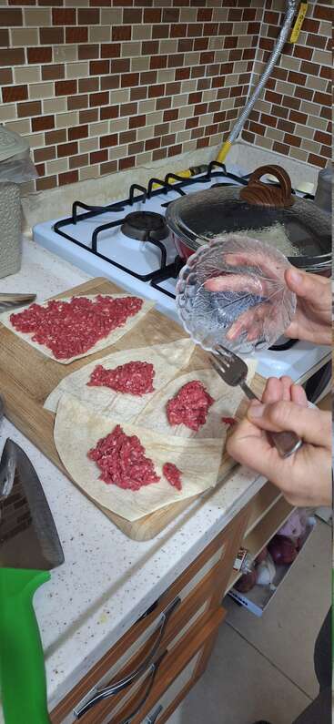 Jemand macht Essen in der Küche und verteilt Hackfleisch auf Fladenbrot auf einem Holzbrett. In der Nähe sieht man Küchenutensilien und einen Herd.