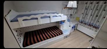 Das ist ein ordentliches Kinderzimmer mit einem weißen Etagenbett, einer gestreiften unteren Matratze, einem kompakten Schreibtisch, Regalen, Büchern, blauen Akzenten und einem Fenster mit dekorativen Vorhängen.