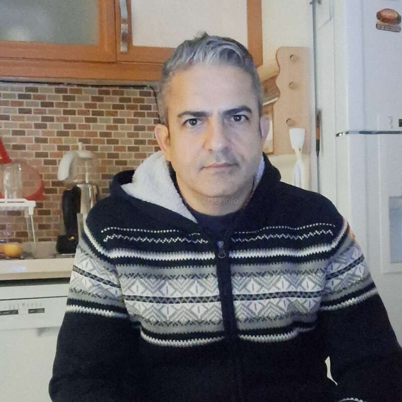 Un homme d'âge moyen aux cheveux gris courts est assis dans une cuisine, vêtu d'un pull à motifs et à fermeture éclair. Il regarde directement la caméra, l'air sérieux.