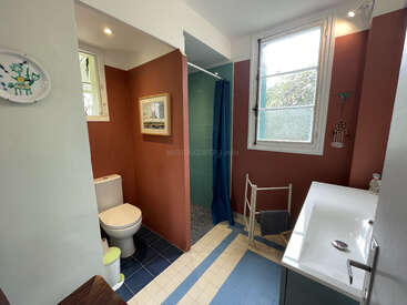 Cette salle de bains présente des murs en terre cuite, des équipements blancs, une douche carrelée de bleu avec un rideau, de grandes fenêtres, un lavabo blanc, un porte-serviettes et une plaque murale décorative. Elle est très lumineuse.