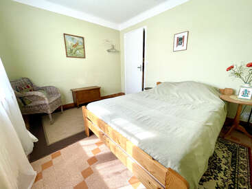 Cette chambre chaleureuse est agrémentée d'un lit en bois, d'un fauteuil en osier, d'une petite table ornée de fleurs, de murs vert clair, de tableaux encadrés, d'un tapis et d'une douce lumière naturelle.