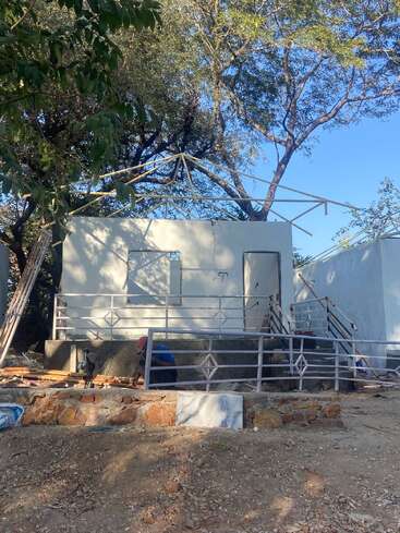 A partially constructed small building with white walls and no roof, surrounded by trees. Construction materials and workers are visible near the structure’s entrance. Sunny day.