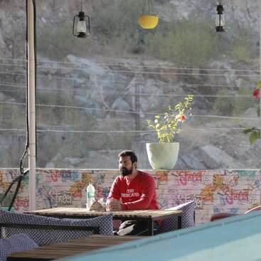 A man in a red sweatshirt sits alone at an outdoor café. Graffiti decorates the walls. Plants, hanging lamps, and mountains fill the background, creating a relaxed atmosphere.