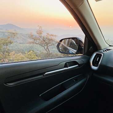 The image shows a serene sunset view of hills and trees, seen through the open window of a parked car’s front seat, creating a peaceful ambiance.