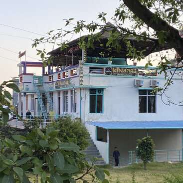 La imagen muestra un edificio blanco con tejado y escalera azules, un cartel que dice "Kinaara" y una rama de árbol en primer plano.