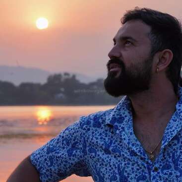 The image depicts a man with dark hair and a beard, wearing a blue and white patterned shirt, standing in front of a serene sunset over a body of water.