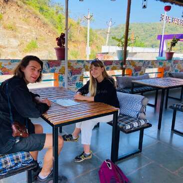 L'image représente un espace extérieur serein avec deux personnes assises à une table, entourées d'un paysage pittoresque de collines et d'arbres sous un ciel bleu clair.