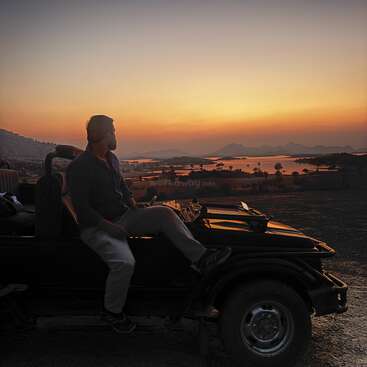 Un hombre está sentado en un jeep aparcado, contemplando una impresionante puesta de sol sobre un lago sereno y montañas lejanas, envuelto en la tranquila luz dorada y anaranjada del atardecer.