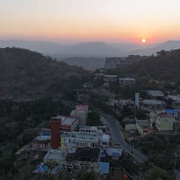 L'image capture un coucher de soleil serein sur une petite ville nichée au milieu des collines. De douces teintes orangées envahissent le ciel, tandis que des routes sinueuses relient les charmants bâtiments colorés en contrebas.