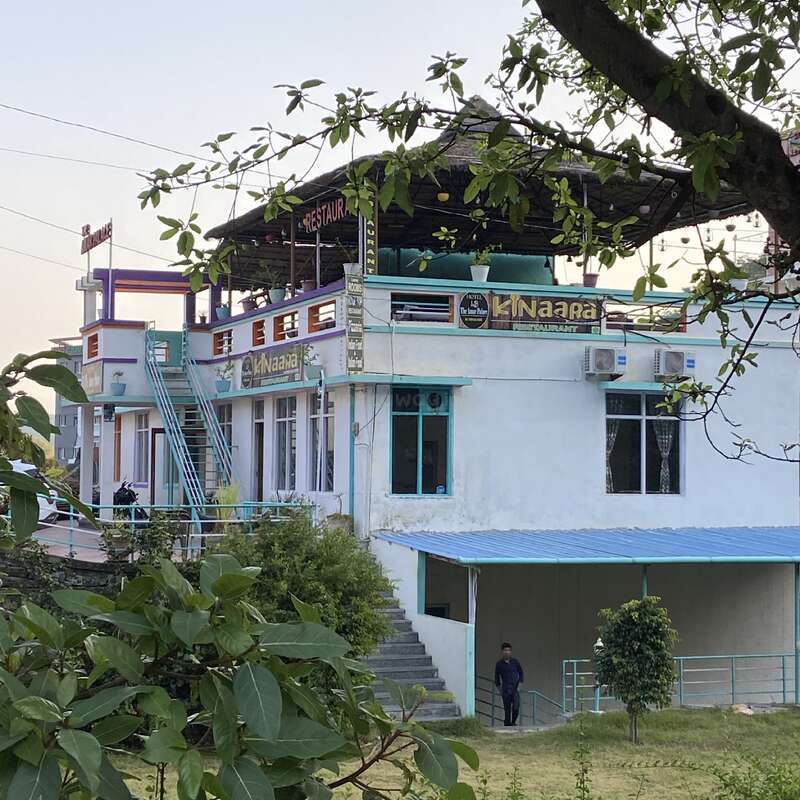 L'image représente un bâtiment blanc avec un toit et un escalier bleus, avec un panneau indiquant "Kinaara" et une branche d'arbre au premier plan.