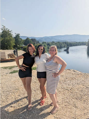 Three women pose happily by a serene river, with mountains and greenery in the background. It’s a sunny day, and they’re dressed casually, enjoying outdoor fun.