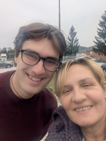 A young man with glasses and a woman are smiling for a selfie outdoors. Trees, cars, and buildings appear in the background on a cloudy day.