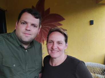 A man and woman smile together indoors. The background is a yellow wall with a large red flower painted on it. Both appear happy and relaxed.