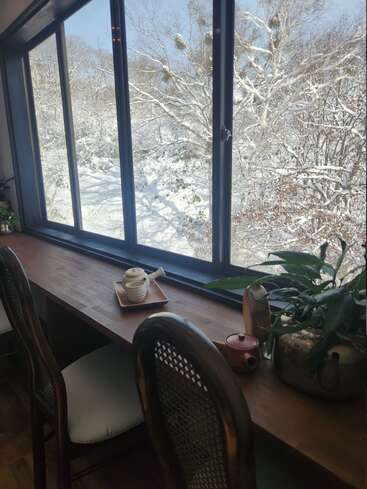 A serene winter scene is visible through a large window. Inside, there’s a wooden counter with teapots, cups, plants, and two chairs, inviting relaxation.