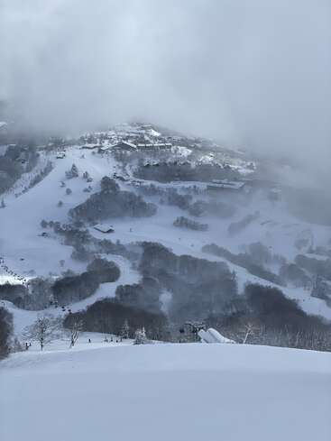 A snowy mountain slope dotted with trees and ski lifts, with a small village nestled in the background, partially covered by mist and clouds, serene winter scene.