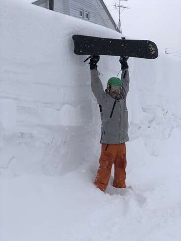 A snowboarder stands knee-deep in snow, holding a snowboard overhead. A massive snow wall and house appear behind, emphasizing the heavy winter conditions and deep snow accumulation.
