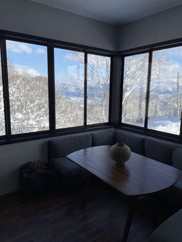 A cozy corner features a modern wooden table with a round vase, surrounded by windows displaying a stunning snowy landscape and distant mountains under a blue sky.