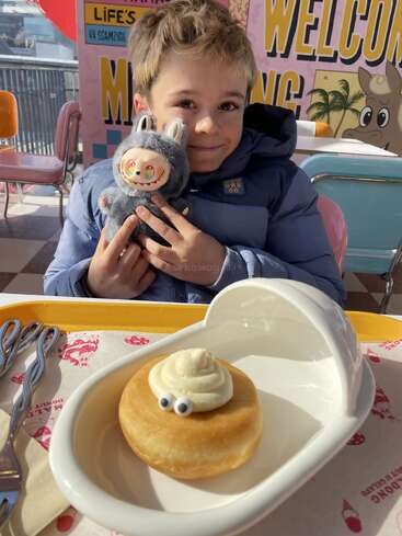 A smiling child in a blue jacket holds a cute stuffed toy. In front, a doughnut with cream and candy eyes sits in a playful dish.