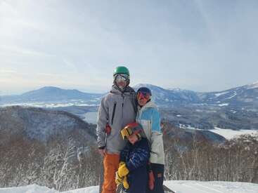 A family of three in colorful ski gear poses on a snowy mountain, surrounded by a beautiful winter landscape with distant hills and a clear, blue sky.