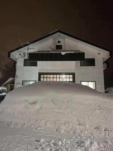 A house stands at night, illuminated windows glowing warmly. A massive mound of freshly fallen snow covers much of the front yard, nearly reaching the windows.