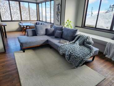 A cozy living room features a large gray sectional sofa, chunky knit blanket, big windows with snowy outdoor views, soft rug, and a minimal, modern design.